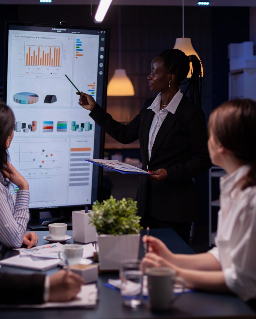 Workaholic african american leader standing in front of presentation monitor explaining marketing project solution late at night in company meeting room. Diverse teamwork working at business ideas.